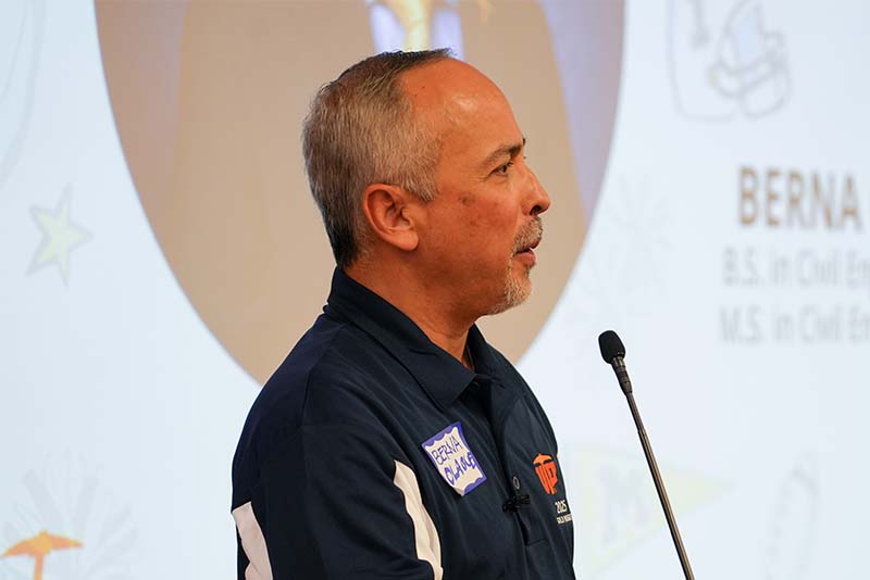 Individual standing near a podium shakes hands with another attendee during a UTEP Engineering homecoming recognition event, with a large projected portrait displayed on the screen behind and attendees gathered in the presentation area.